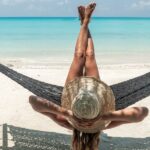Eine Frau entspannt in einer Hängematte zwischen zwei Palmen und blickt auf das Meer. Sie trägt einen Strohhut und genießt die Sonne, was ihr hilft, die Eigenschutzzeit der Haut zu verlängern. Ihre überkreuzten Beine und die tropische Atmosphäre vermitteln Ruhe und Entspannung unter der Sonne.